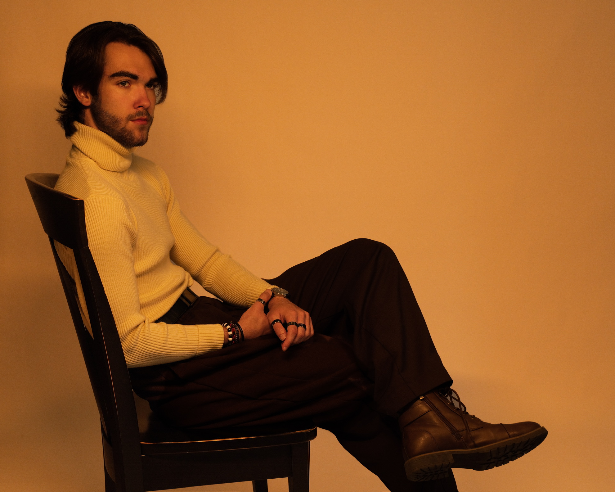 Richard Jordan seated in a chair wearing a cream turtleneck and dark trousers against a warm amber backdrop
